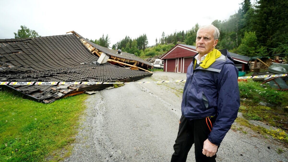 Jonas Gahr Støre står foran en kollapset bygning med et skadet tak, omgitt av rusk og polititape, i landlige omgivelser.