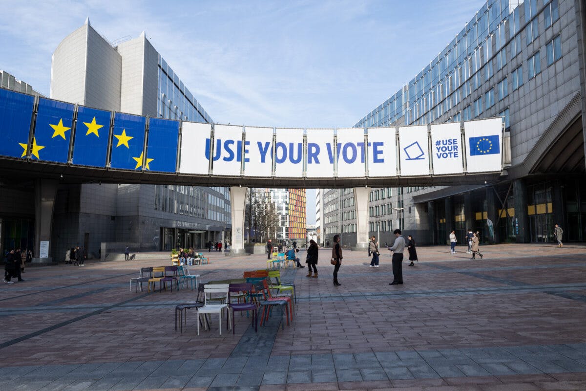 Et offentlig torg med folk som går, med et stort «bruk din stemme»-banner med eu-stjerner, flankert av moderne bygninger.