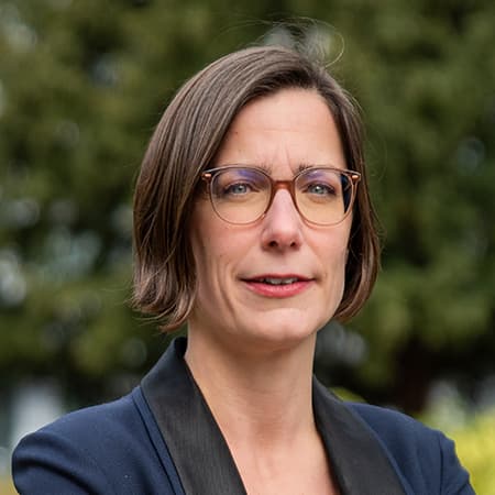 En kvinne med kort brunt hår og briller iført en mørk blazer står utendørs foran grøntområder og ser inn i kameraet.