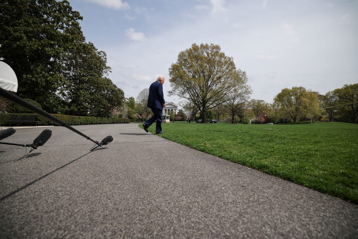 En person i dress går alene på en asfaltert sti gjennom et gresskledd område med trær og mikrofoner synlige i forgrunnen.