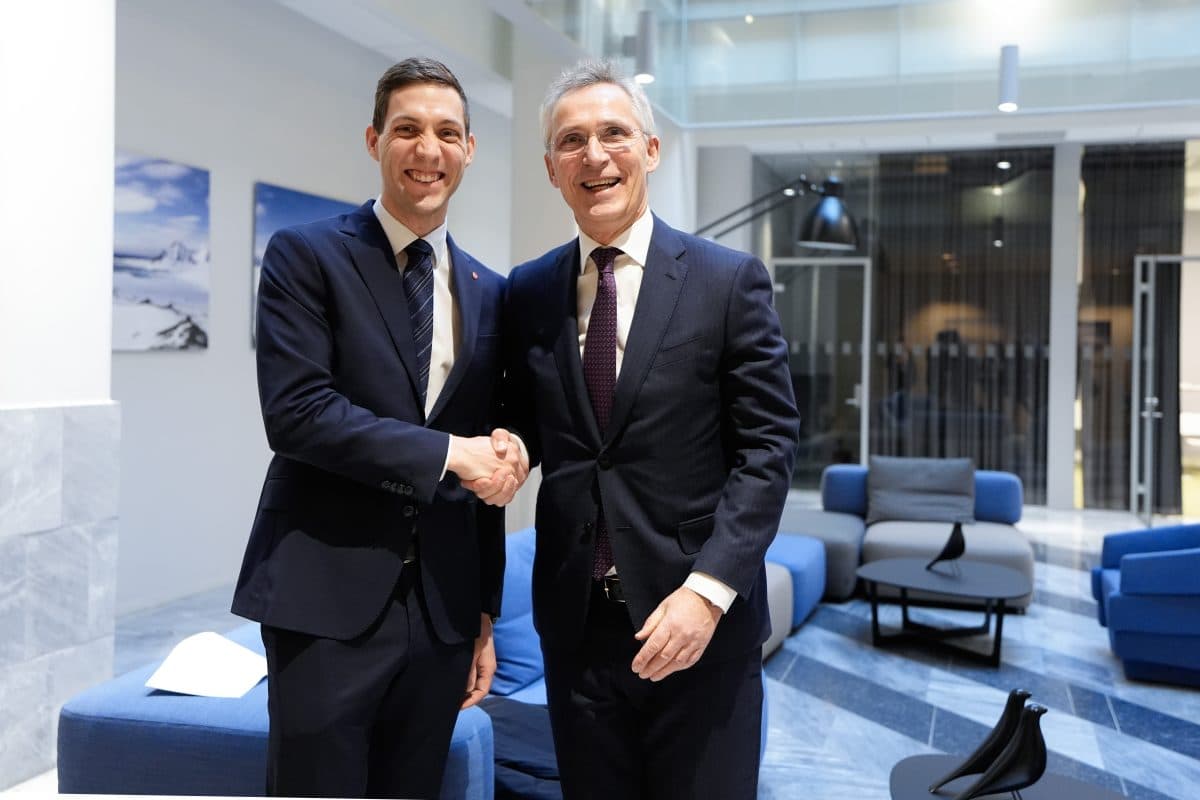 To menn i dress håndhilser og smiler i en moderne kontorlobby med blå møbler og store vinduer.