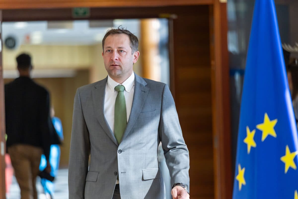 En mann i grå dress og grønt slips går innendørs i nærheten av et EU-flagg.