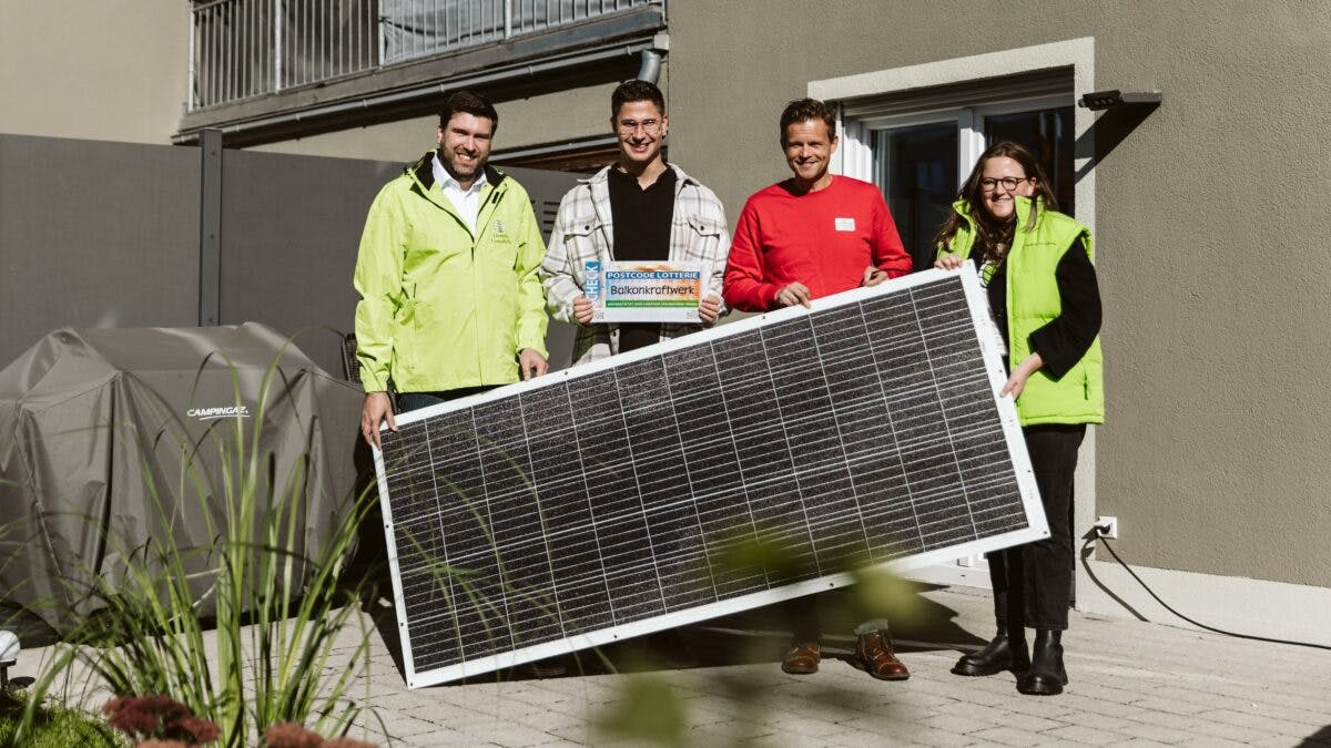 Fire personer står utendørs og holder et stort solcellepanel. De ser ut til å smile. En person holder et lite skilt. Et dekket objekt og bygning er i bakgrunnen.