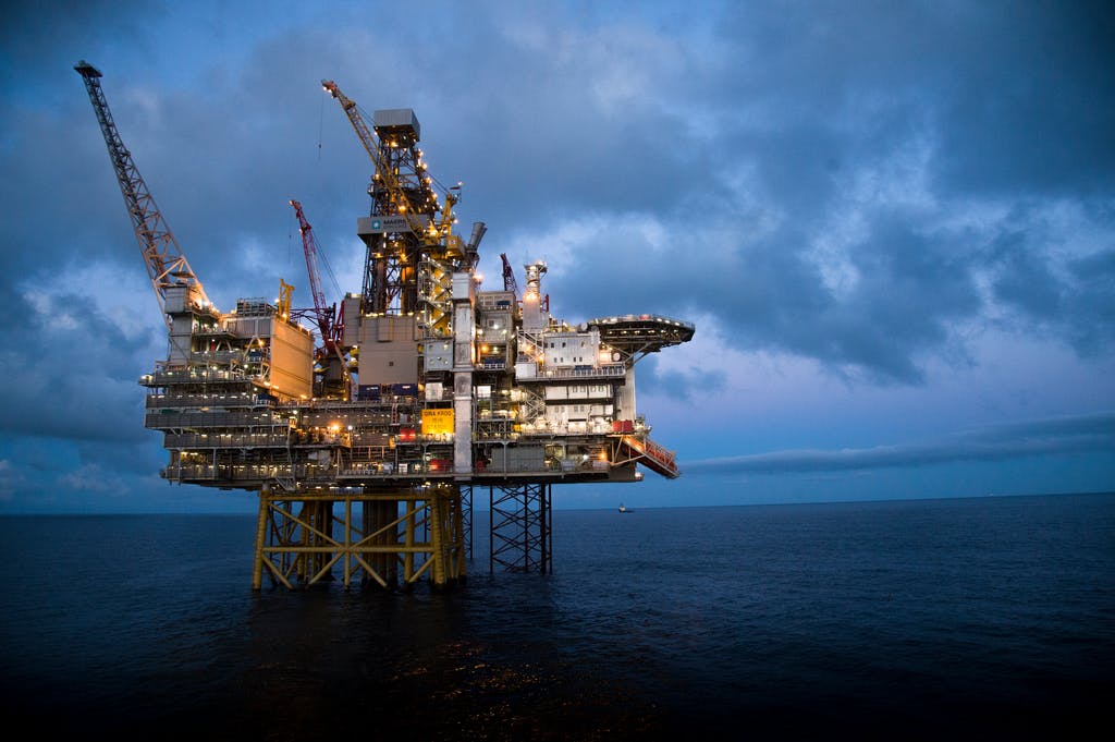 En offshore oljerigg står opplyst mot en overskyet kveldshimmel, med kraner og utstyr synlig, plassert midt i havet.