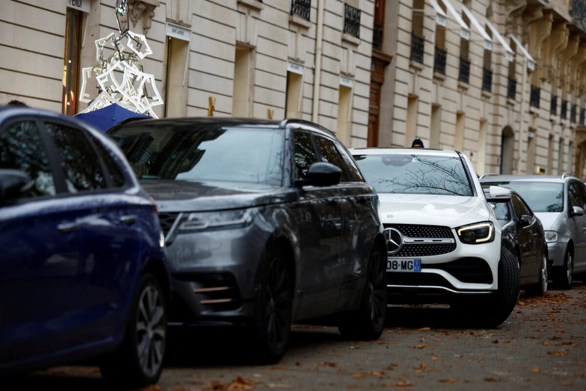 Flere biler, inkludert en hvit Mercedes og en grå SUV, står parkert langs siden av en bygate med bygninger.