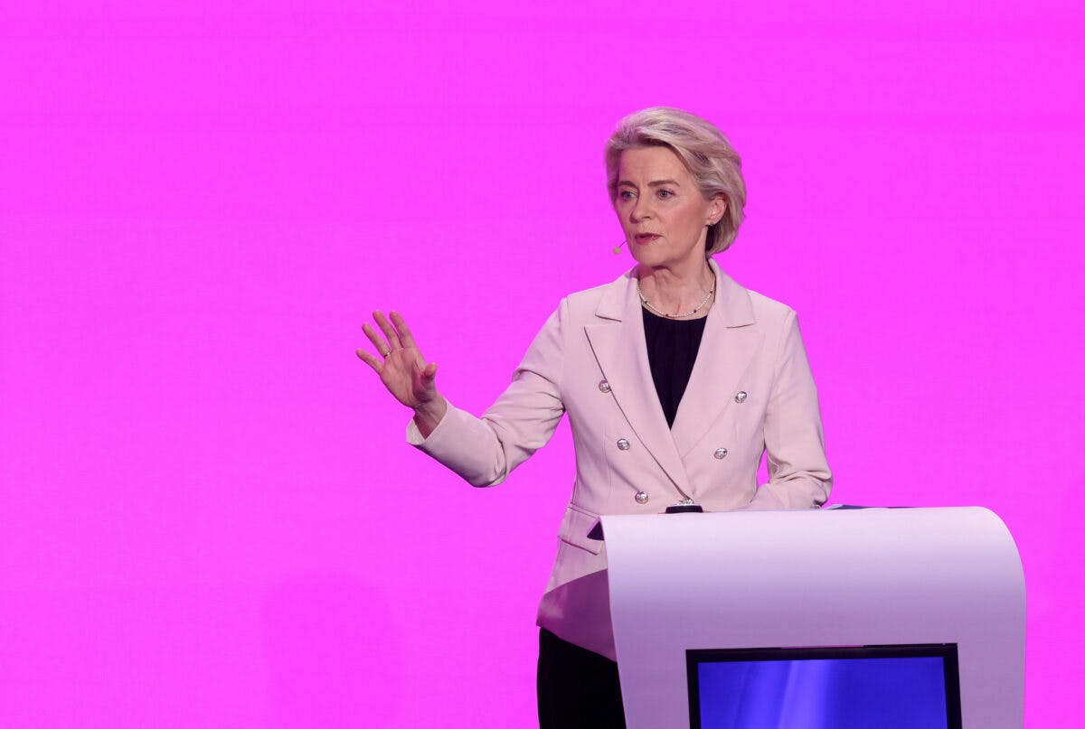 En kvinne står bak et hvitt podium og gestikulerer med en hånd. Hun har på seg en lys blazer og snakker mot en knallrosa bakgrunn.