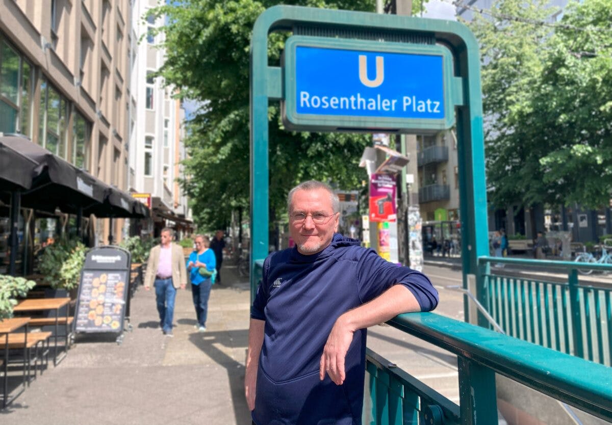 En mann i en blå hettegenser står ved siden av et skilt til Rosenthaler Platz på en solrik dag. Butikker, mennesker og uteservering er synlige i bakgrunnen.