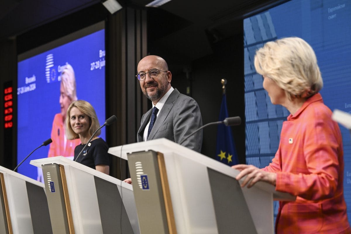 Tre personer på pallene deltar på en pressekonferanse foran et EU-bakteppe. To av dem ser på mannen i midten mens han snakker.