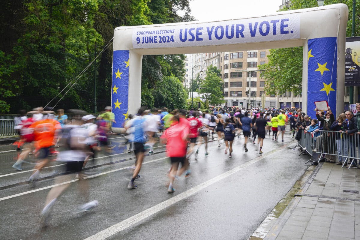 En gruppe løpere i bevegelse passerer under en stor oppblåsbar buegang med teksten "European Elections 9 June 2024 - Use Your Vote," på en regnfull vei. Tilskuere står bak barrierer.