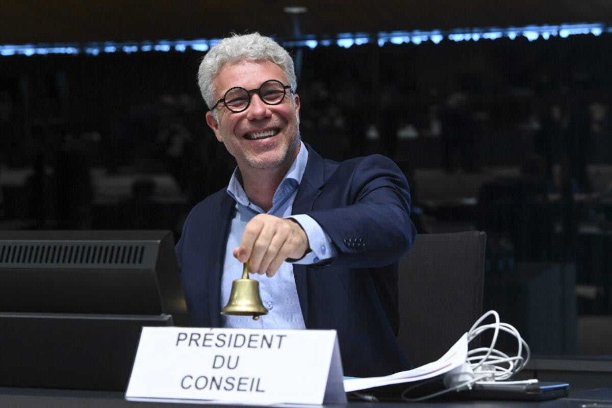 En mann med briller, smilende, holder en liten bjelle i hånden mens han sitter ved et skrivebord med et skilt som leser «President du Conseil.