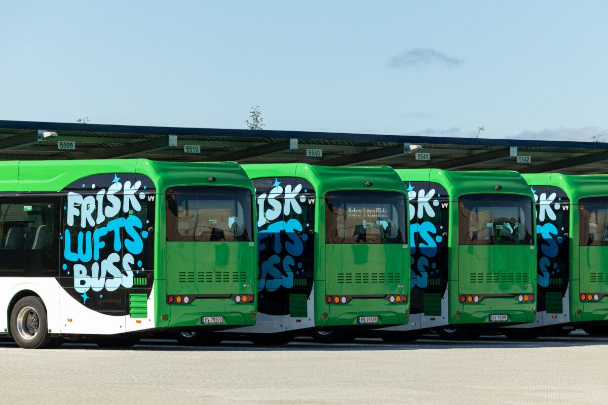 Seks grønne busser er parkert på rad ved en bussterminal under overskyet himmel, med ordene "FRISK LUFTS BUSS" malt tydelig på sidene.