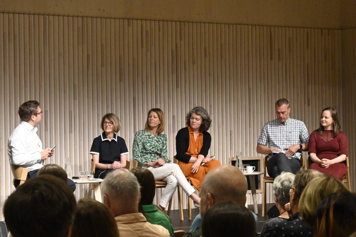 Et panel på fem personer sitter på scenen foran et publikum. Fire kvinner og en mann diskuterer mens en sjette person modererer diskusjonen. De er i et rom med trepanel.