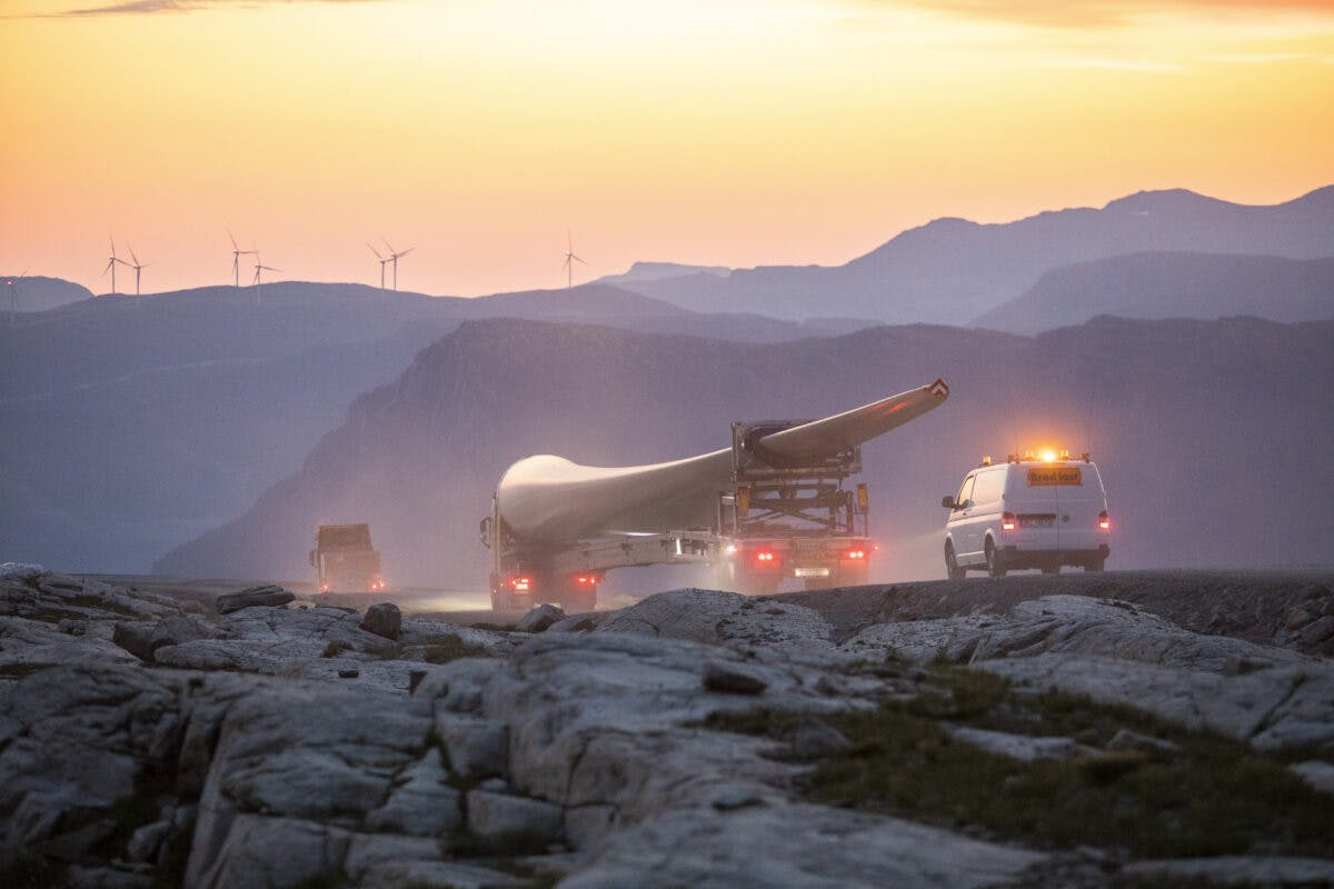 En lastebil frakter et vindturbinblad på en fjellvei ved solnedgang, etterfulgt av en hvit varebil med blinkende lys. Vindturbiner og fjelltopper er synlige i bakgrunnen.