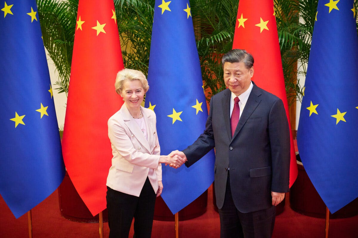To personer håndhilser og står foran EU- og kinesiske flagg. Den ene er i lys dress, den andre i mørk dress. Grønne planter er i bakgrunnen.