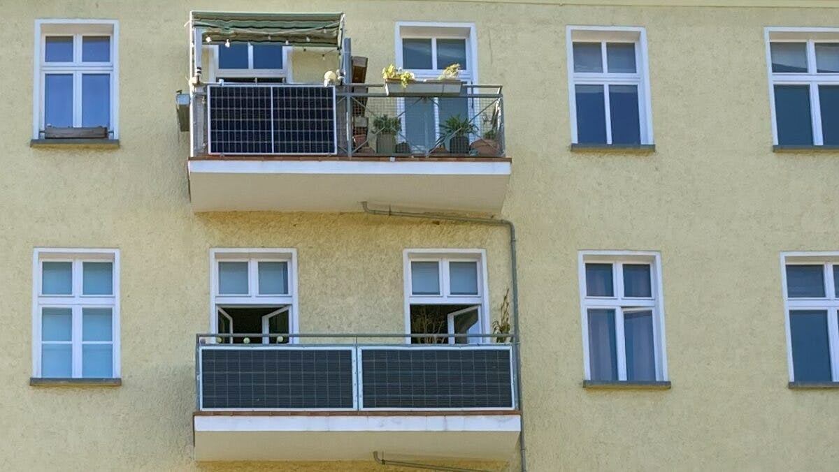 To balkonger på et lysegult bolighus med solcellepaneler, potteplanter, og en markise på øvre balkong.