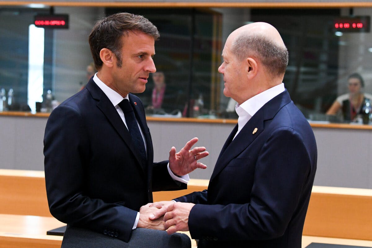 To menn i dress snakker og håndhilser i et moderne møterom. Den ene gestikulerer med hånden mens den andre lytter oppmerksomt.
