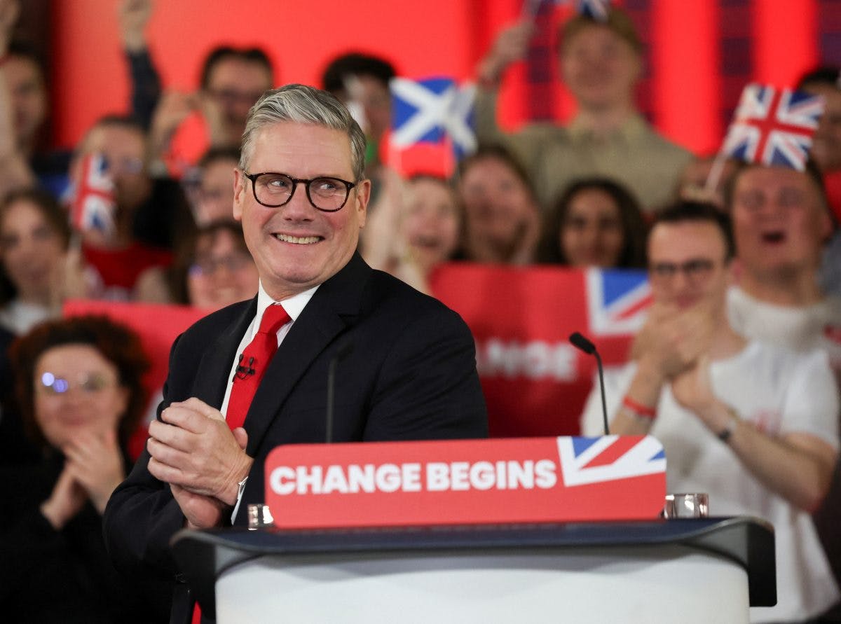 En mann i dress, som står på et podium med et «CHANGE BEGINS»-skilt, klapper med folk i bakgrunnen som holder britiske og skotske flagg.
