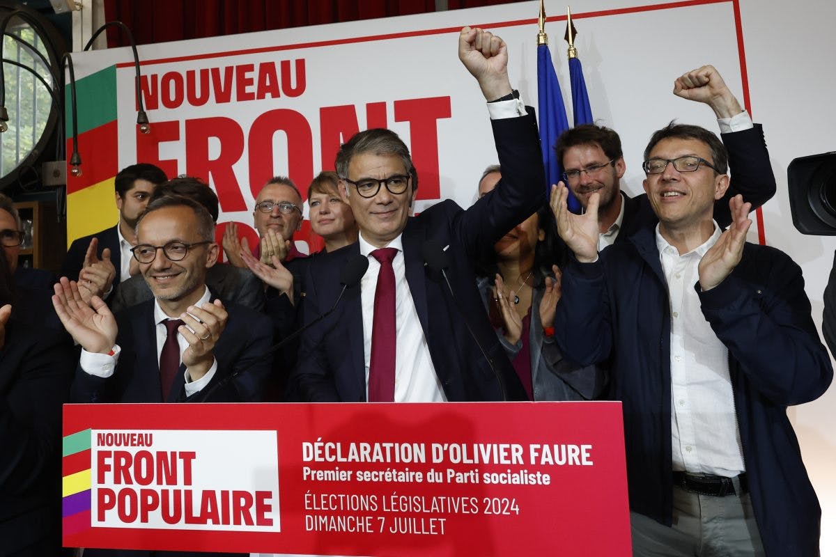 En mann med briller snakker ved et podium med «Nouveau Front Populaire» i bakgrunnen, flankert av applauderende folk, noen heve knyttnever.