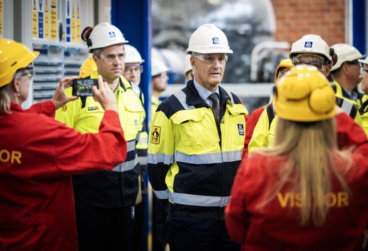 En gruppe mennesker i vernehjelmer og synlige jakker på turné i et industrianlegg. Noen individer bærer røde jakker merket "VISITOR", mens andre bruker gule jakker og hjelmer.