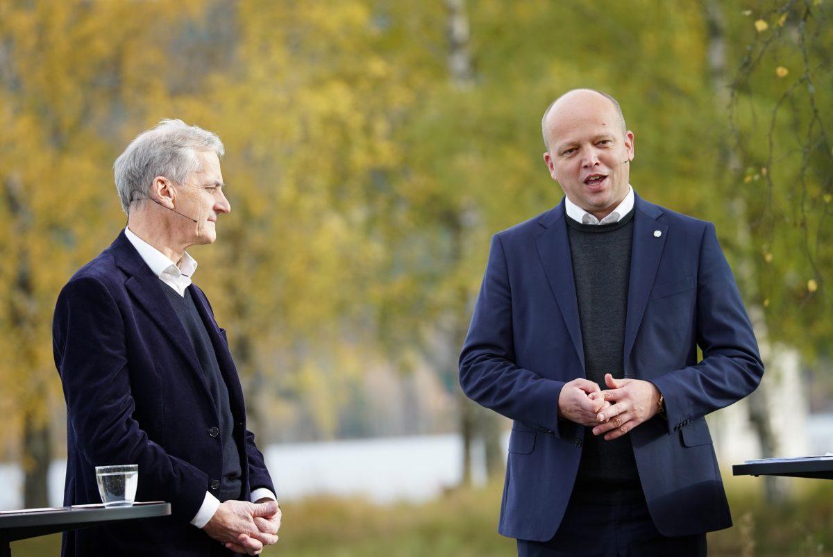 To menn i formell antrekk står utendørs og er i samtale. En mann snakker mens den andre lytter. Bakgrunnen har grønt og høstløvverk.