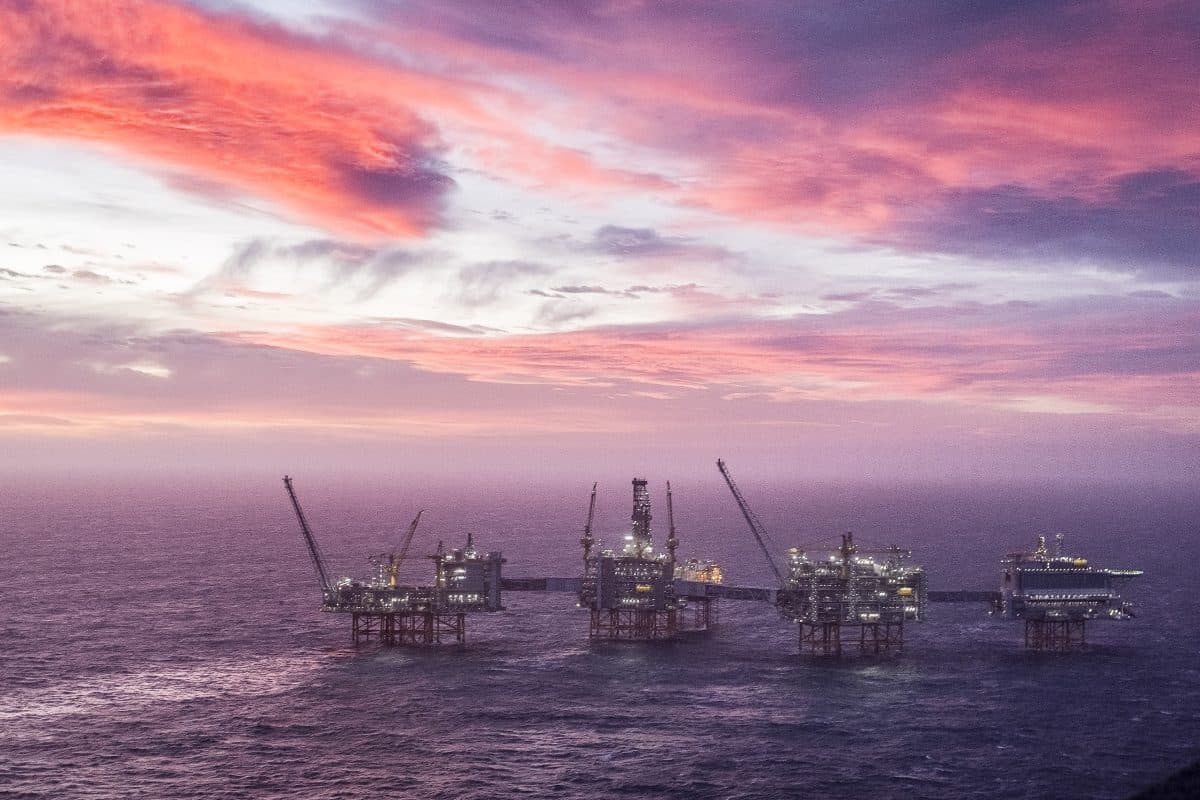 Et oljeplattformkompleks til havs er satt mot en fargerik solnedgangshimmel, med flere strukturer forbundet over havvannet.