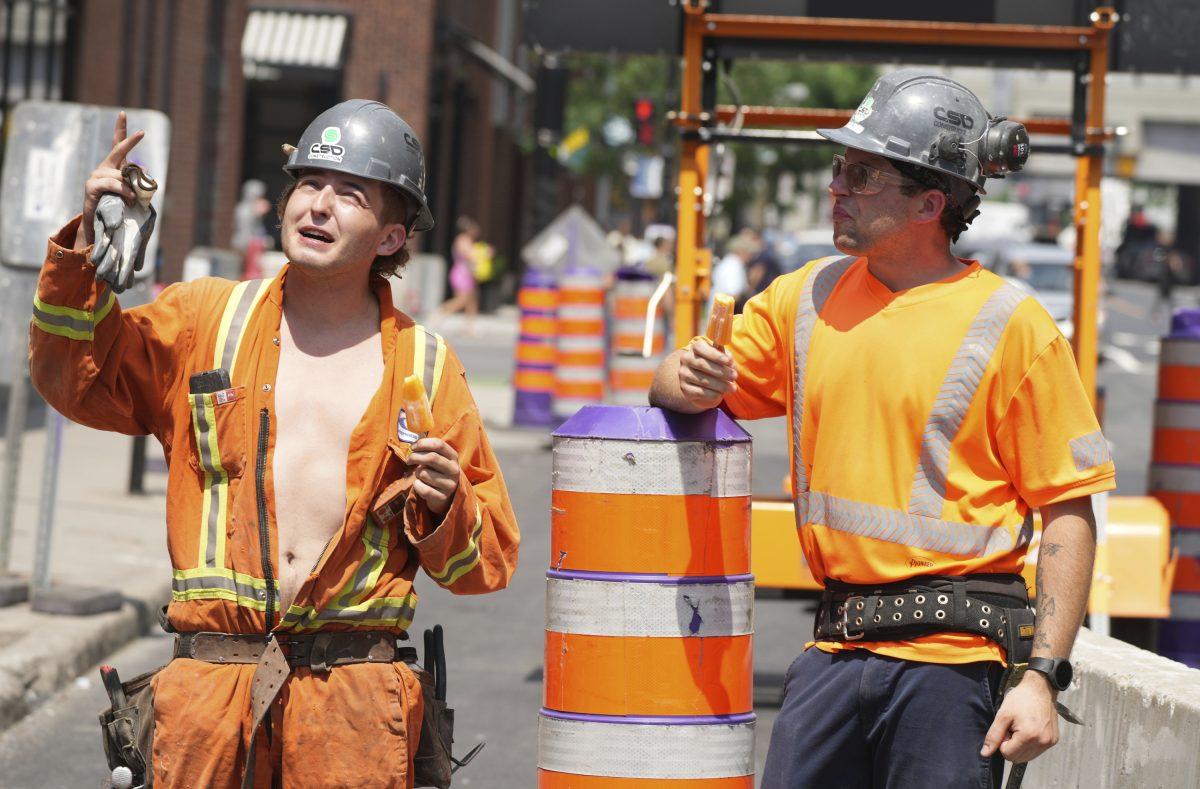 To bygningsarbeidere i verneutstyr snakker sammen på en byggeplass. Den ene arbeideren gestikulerer oppover mens han holder en hanske, og den andre lytter og lener seg på en oransje trafikktønne.