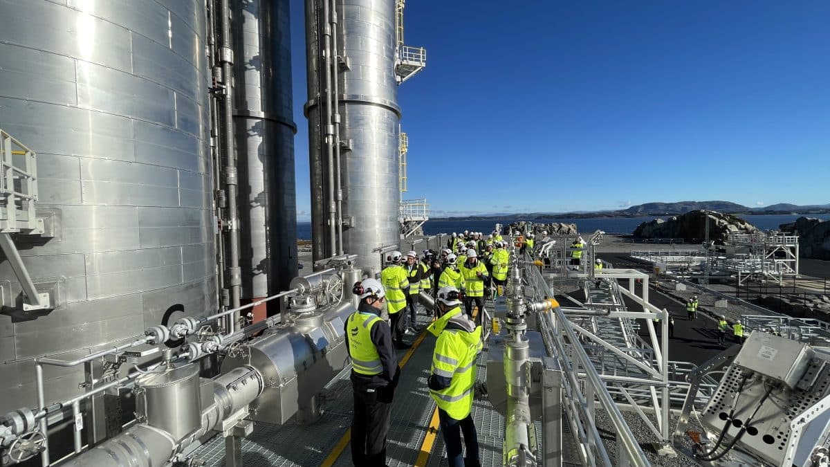 En gruppe mennesker i sikkerhetsutstyr med høy synlighet besøker et industrianlegg med store metallkonstruksjoner og rør. Bakgrunnen viser en klar himmel og et fjernt landskap.