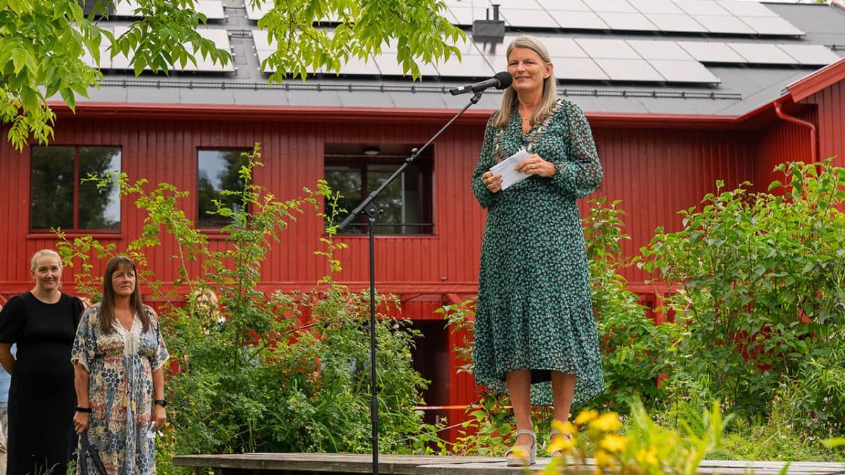 En kvinne står på en forhøyet treplattform og snakker inn i en mikrofon. Hun står foran et rødt bygg med solcellepaneler på taket, omgitt av grøntområder. To andre kvinner står i nærheten.