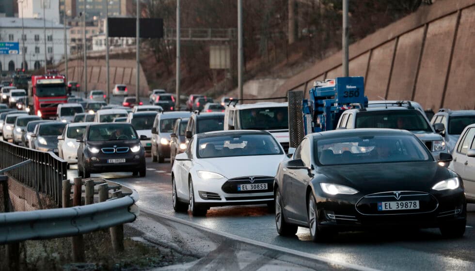 Trafikkbelastning på en flerfeltsvei med biler og lastebiler i begge retninger, avgrenset av bommer og bybebyggelse i bakgrunnen.