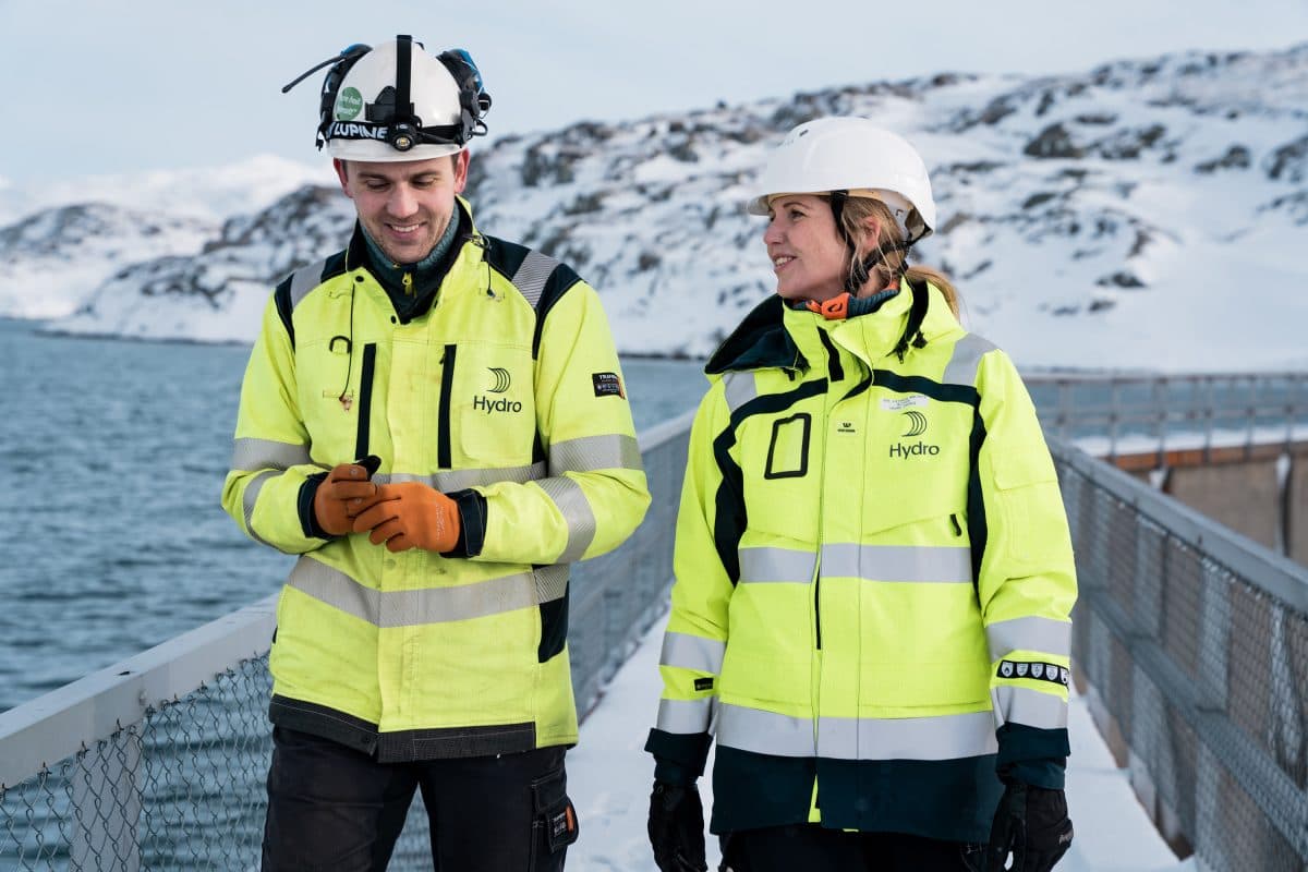 To personer i gule vernevester og hjelmer står på en snødekt sti ved en vannmasse, med steinete, snødekte åser i bakgrunnen.