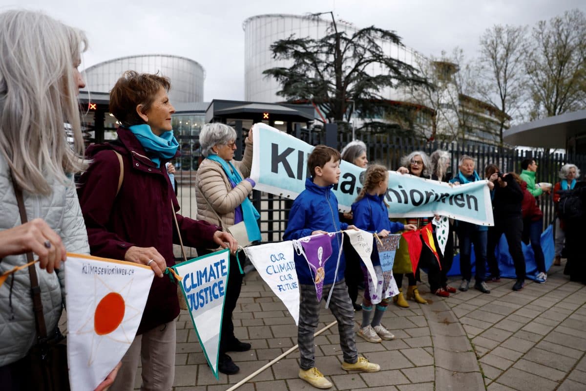 En gruppe mennesker står i kø og holder skilt og bannere som tar til orde for klimarettferdighet utenfor en stor moderne bygning.