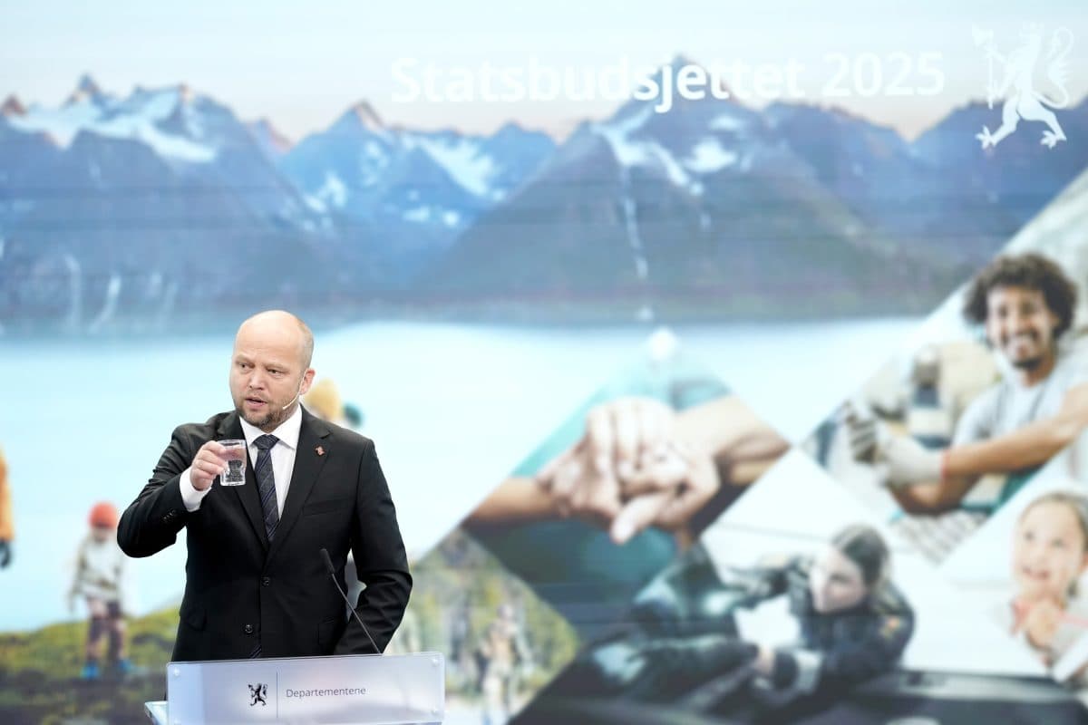 En person som snakker på et podium foran et bakteppe med fjell og ulike livsstilsbilder, med teksten "Statsbudsjettet 2025" vist over.