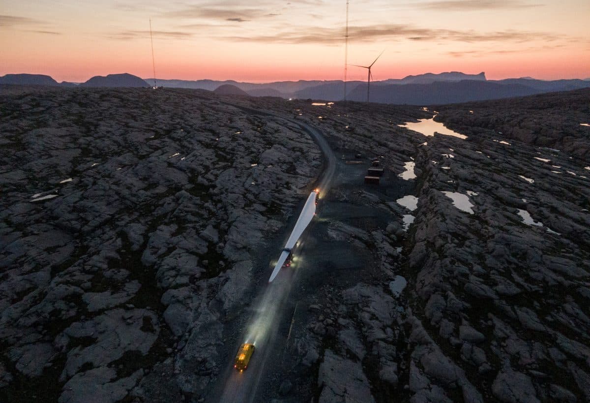 Luftfoto av en lastebil som frakter et langt vindturbinblad langs en svingete vei med et steinete landskap ved solnedgang, med vindturbiner i bakgrunnen.