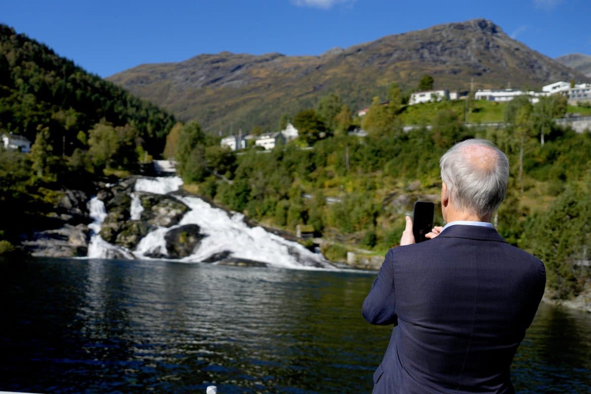 En mann i dress tar et bilde av en foss og et fjellandskap.