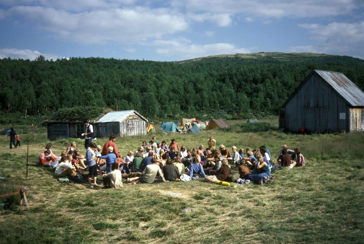 En gruppe mennesker som sitter i en sirkel på gress i et landlig område, med telt og trebygninger i bakgrunnen.