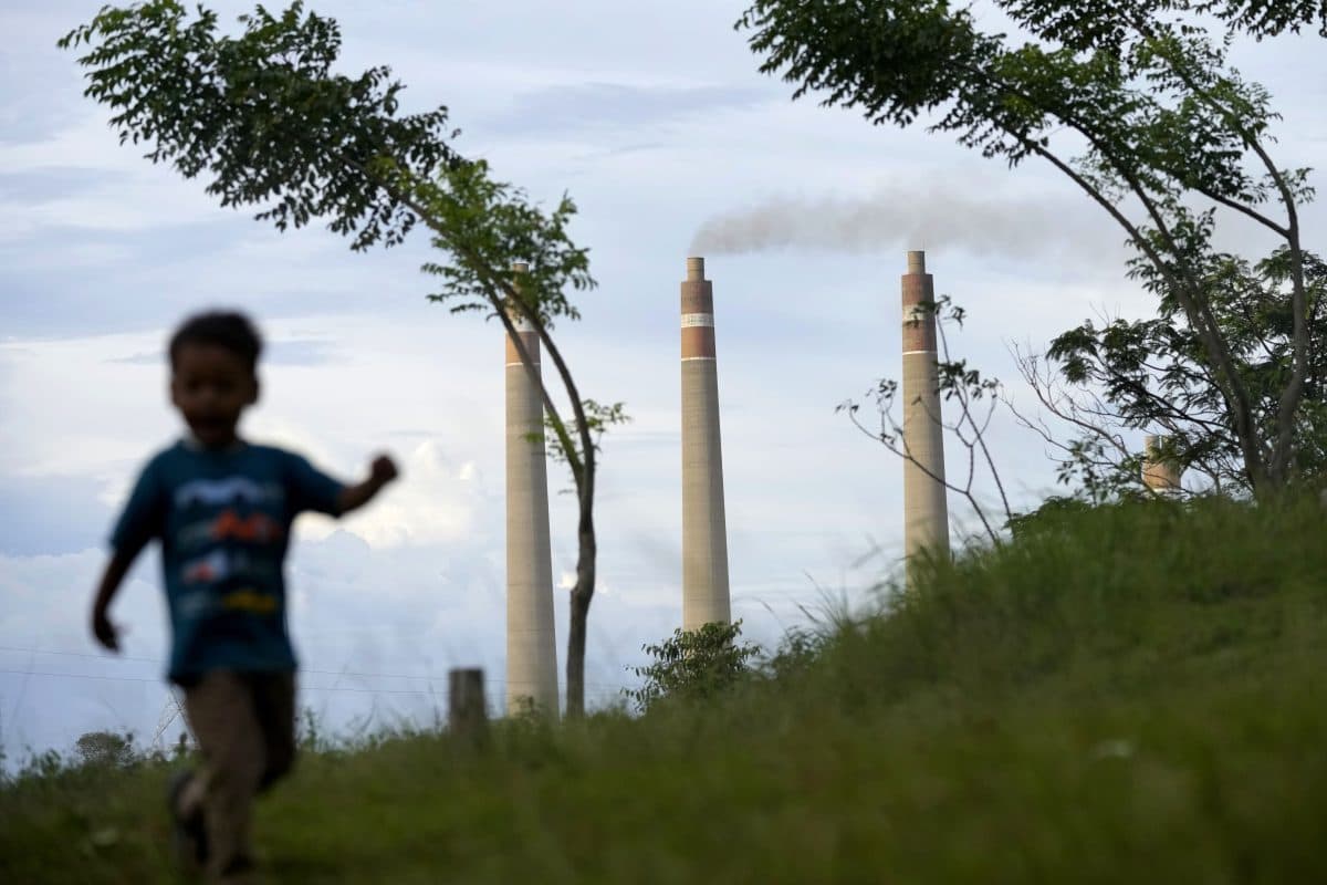 Et barn løper på gress i forgrunnen, med to rykende industrielle skorsteiner i bakgrunnen mot en overskyet himmel.