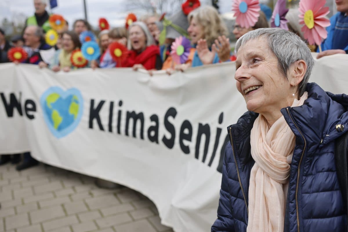 En smilende eldre kvinne står foran en gruppe og holder et banner med teksten "Vi [hjerte] KlimaSeniorinnen", med fargerike blomster.