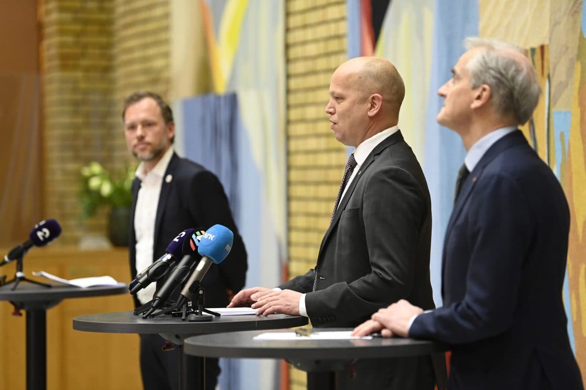 Tre menn i dress står ved podier med mikrofoner under en pressekonferanse i et rom med fargerike vegger.