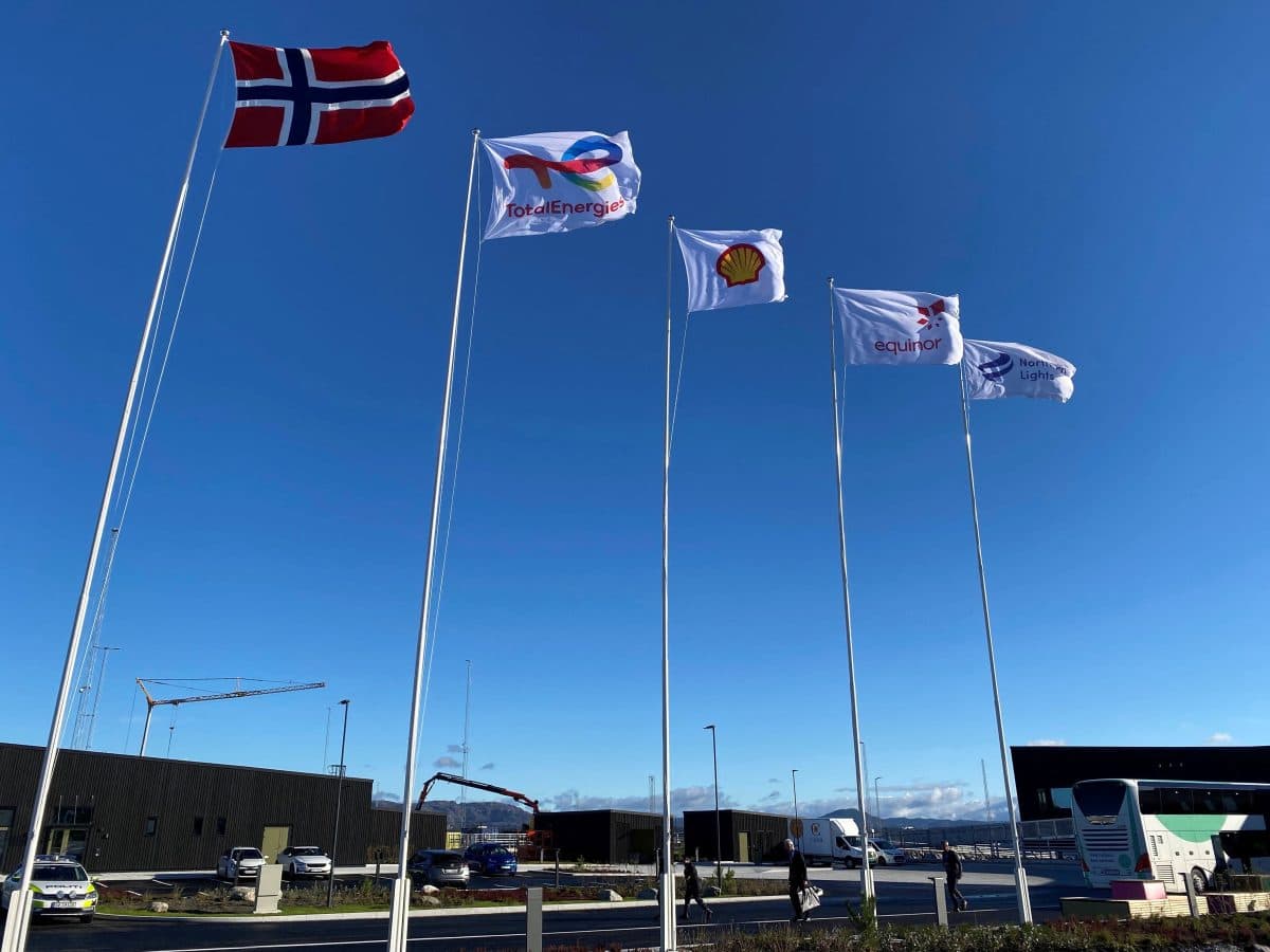 Fem flagg på stenger, inkludert det norske flagget og flagg fra energiselskaper, vaier i vinden mot en klarblå himmel med bygninger og kjøretøy synlige i bakgrunnen.