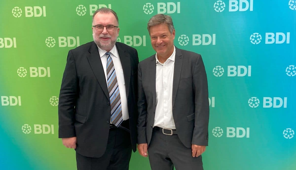 To menn i dress står foran en grønn og blå bakgrunn med «BDI»-logoer. De smiler til kameraet.