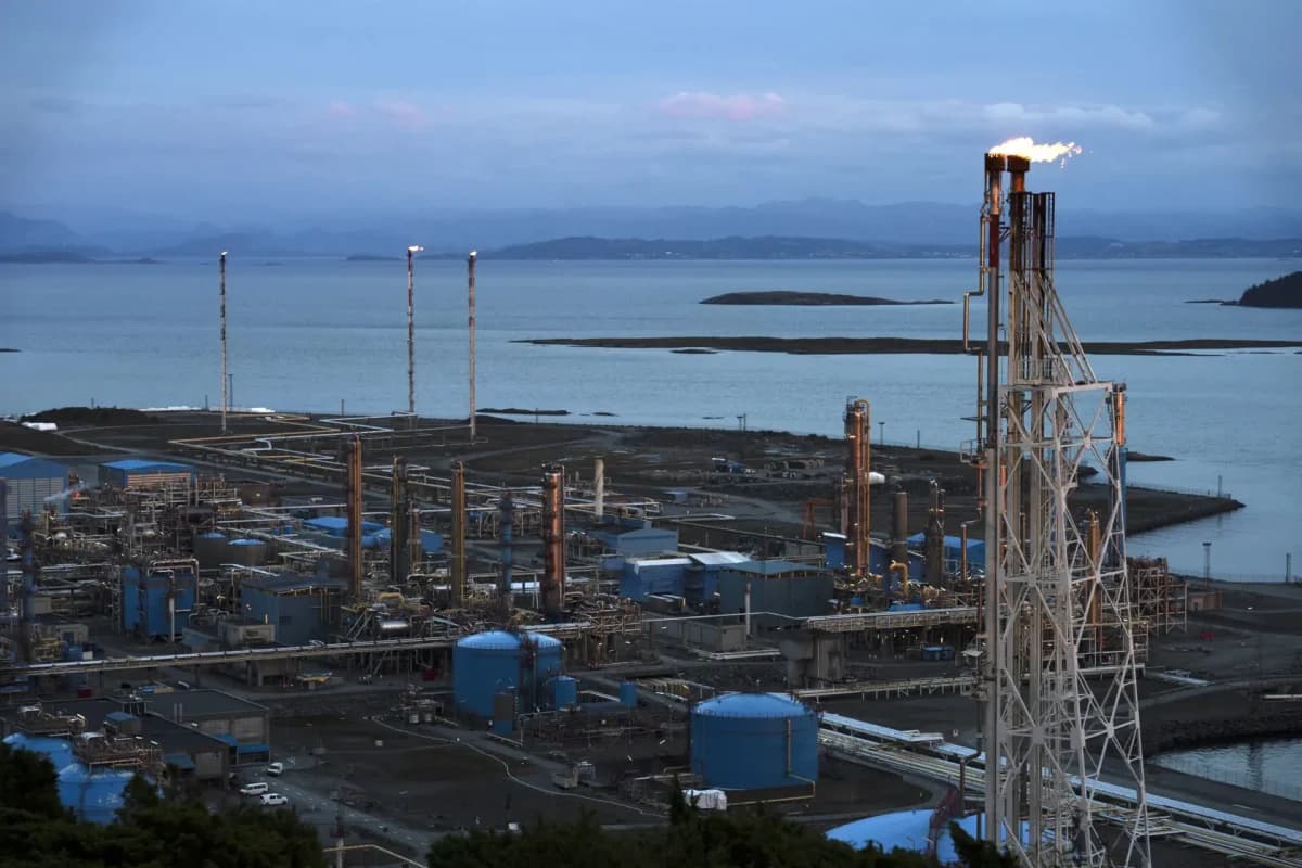 Et kystindustrielt raffineri med flere skorsteiner og lagringstanker, satt mot et bakteppe av vann og fjerne åser under en overskyet himmel.