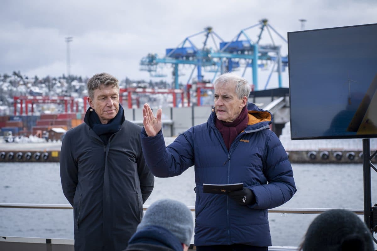 To menn står utendørs ved en vannkant. En snakker mens han holder et hefte. Fraktkraner og containere er synlige i bakgrunnen. Det ser ut til å være en kald dag.