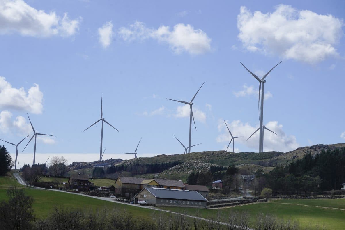 Vindturbiner i en grønn åsside med blå himmel og spredte skyer. Gårdsbygninger er synlige i forgrunnen.