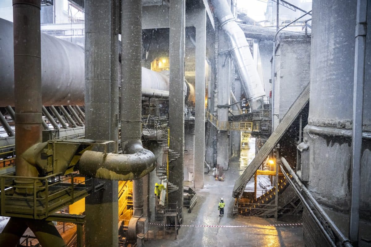 Innvendig visning av et industrianlegg med store rør, metallkonstruksjoner og noen få arbeidere i sikkerhetsutstyr.