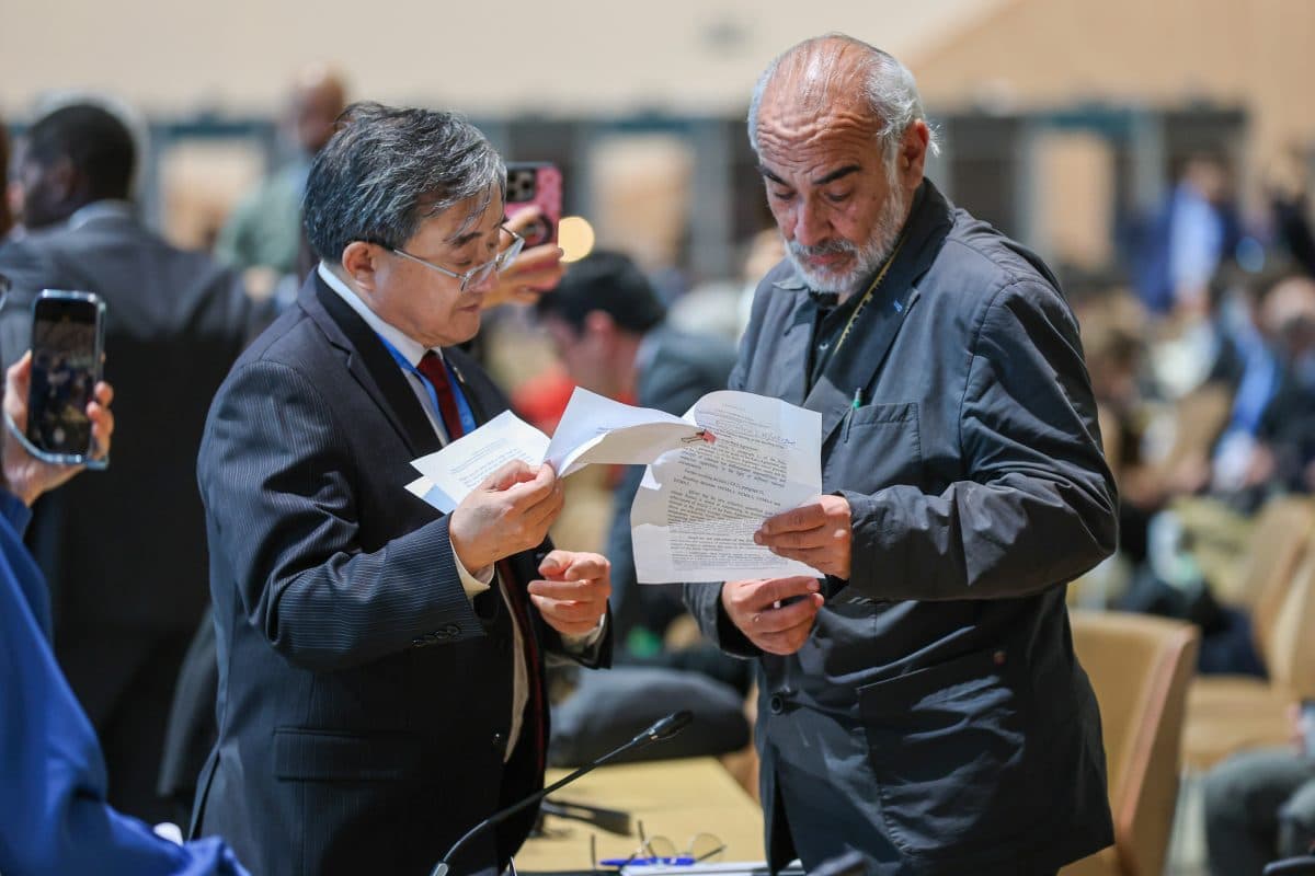 To menn diskuterer dokumenter i et konferanserom. Den ene mannen gestikulerer med et papir mens den andre lytter oppmerksomt. Publikumsmedlemmer og konferanseplasser er synlige i bakgrunnen.