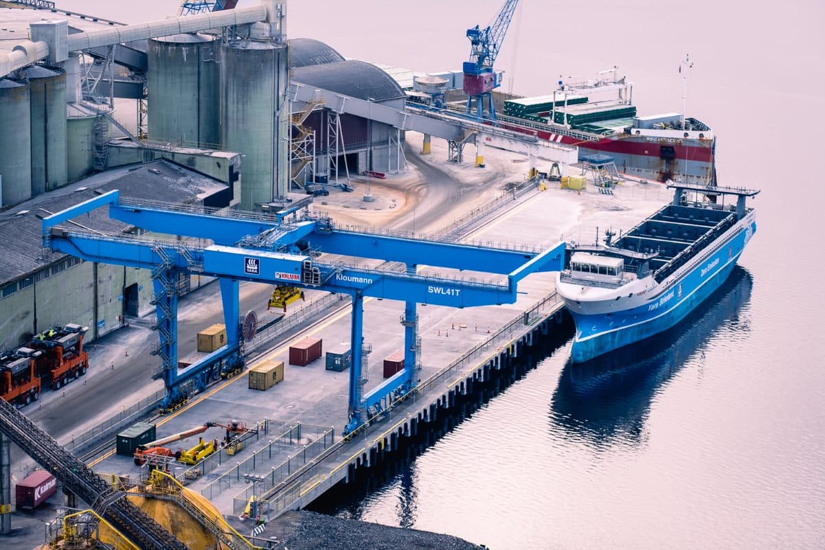 Et forankret lasteskip ved en industrihavn med kraner og store lagringsanlegg rundt.