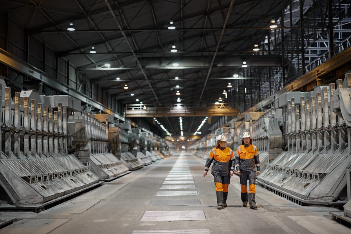 To arbeidere i oransje uniformer går nedover en industrifabrikkgang foret med store maskineri under et høyt tak.