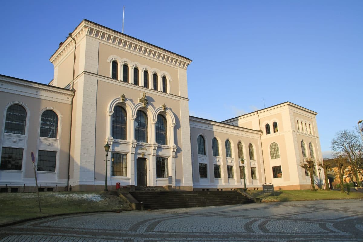 En stor, klassisk bygning med buede vinduer og lys fasade står under en klar blå himmel, med et brosteinsområde foran og et bladløst tre til høyre.