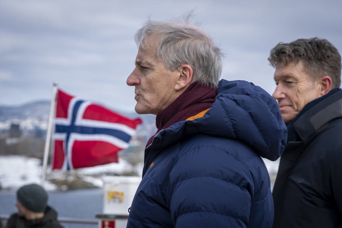 To menn i vinterjakker står utendørs med et norsk flagg i bakgrunnen. Snødekt landskap og overskyet himmel synlig.