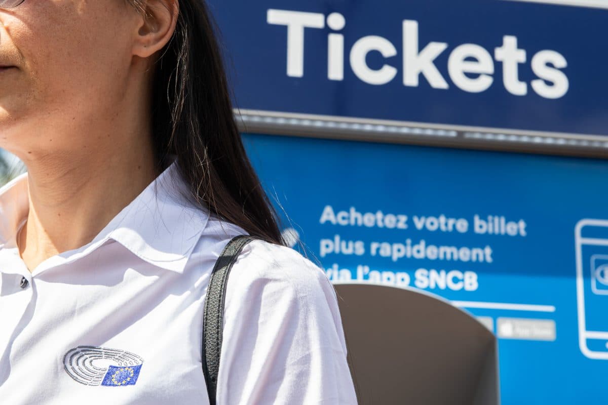 En person iført hvit skjorte står foran en billettautomat og viser et skilt på fransk om å kjøpe billetter via SNCB-appen.